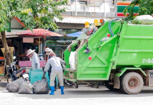 In the foreground, two workers wearing protective gloves, white overalls, and blue rubber boots are collecting rubbish into large black garbage bags placed on the pavement. Behind them, a bright green waste collection truck with a hydraulic lifting mechanism and safety lights is positioned on the street, ready to haul away waste. The truck's rear section is equipped with various tools and hoses used during rubbish collection. To the left of the workers, there are trees with green leaves partially shading the scene, and behind the truck, a small market stall with a pink umbrella and other colourful awnings is visible, suggesting a busy urban area. The setting appears to be outside a retail or market location on a typical street in Enfield, where Enfield House Clearance services may operate, with environmental surroundings including residential or commercial buildings and other street fixtures. The overall scene depicts a professional rubbish collection operation in a suburban town environment with natural daylight illuminating the area.
